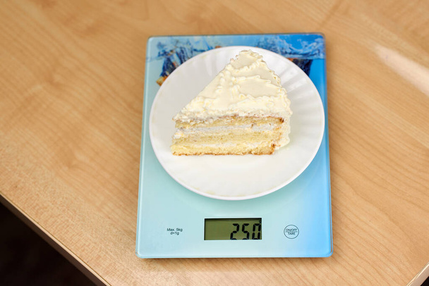 A piece of delicious cake with white butter cream lies on the scales. Calorie counting concept, figure tracking - Photo, Image