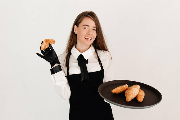 Πορτρέτο κομψής σερβιτόρας με μαύρη ποδιά και γάντια που σερβίρει πελάτες σε ζαχαροπλαστείο, δίνοντας κρουασάν - Φωτογραφία, εικόνα