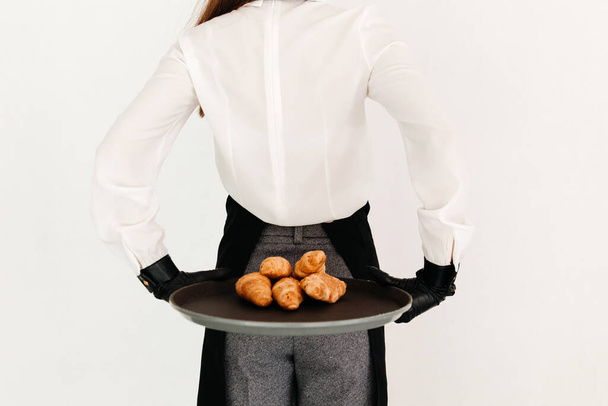 Zdjęcie eleganckiej kelnerki w czarnym fartuchu, trzymającej tacę z rogalikami za plecami w cukierni - Zdjęcie, obraz