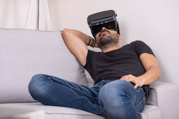 Young man in VR glasses headset at home on sofa , future technology, education video gaming - 写真・画像