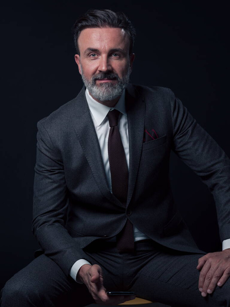 Portrait of adult businessman wearing trendy suit and sitting in modern studio on stylish chair against the black background. Horizontal mockup. High quality photo - Foto, afbeelding