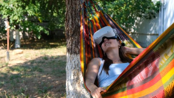 funny woman wearing virtual reality glasses rests in a hammock near a tree, imagining a dream come true, an invented world. The concept of getting experience using VR headset glasses of virtual - Photo, Image