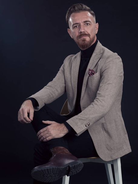 Portrait of adult businessman wearing trendy suit and sitting in modern studio on stylish chair against the black background. Horizontal mockup. High quality photo - Foto, imagen