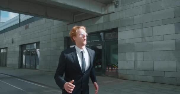 Young businessman runs down city street. Business man late for meeting. Successful man in formal suit jogging near modern office buildings. Red-haired male manager in hurry to appointment. - Footage, Video