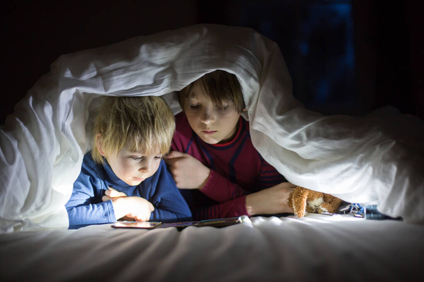 Adorable blond toddler child and older brother, cute boys, reading little book with little toy next to him under the duvet in bed - Fotoğraf, Görsel