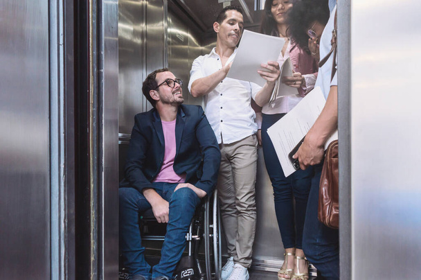 男女の男女が混じってカジュアルな服を着た車椅子の男性が同僚のエレベーターの中で口論していました. - 写真・画像