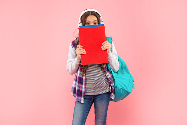 teen girl with backpack behind workbook wearing headphones on pink background, ebook - Valokuva, kuva