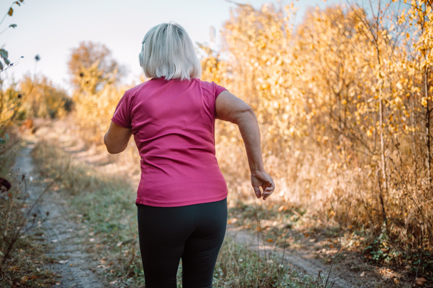 Elderly woman running with headphones and smart watch in a park in a beautiful autumn nature. Nature and sport healthy lifestyle concept. - Photo, image