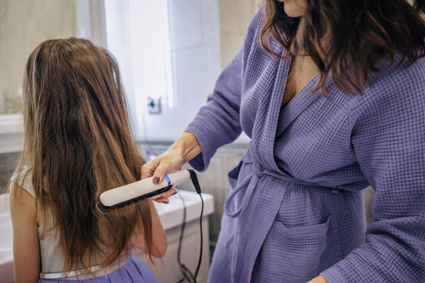 Mom mother ironing hair straightener tongs to smooth hair of daughter to girl. Concept for grooming and hairstyling, preparing for school. - Foto, Imagen