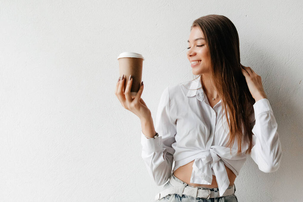 Young brunette woman holding disposable coffee cup and looking at the coffee cup. Morning routine concept - Foto, afbeelding