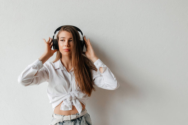 Beautiful young brunette woman in white shirt and jeans posing over white background. Listening to music with headphones - Fotó, kép