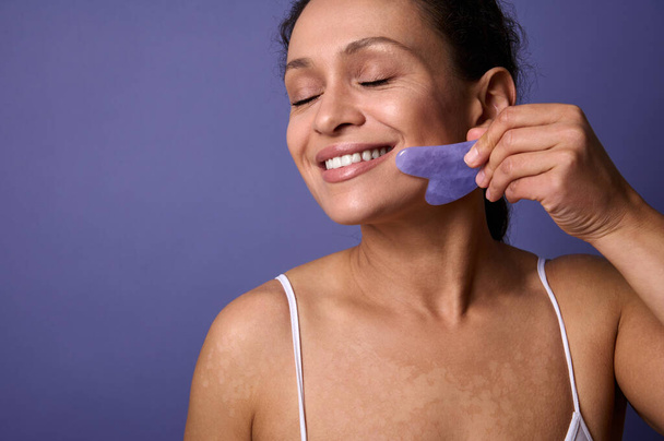 Close-up portrait of delightful self-confident natural beauty woman with vitiligo skin problems smiling toothy smile massaging her face with Ayurvedic Gua-Sha jade massager against violet background - Foto, imagen