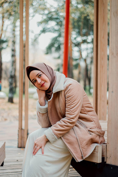Muslim woman in beige coat and headscarf. Stylish Muslim woman in the park. A girl sitting on a bench - Fotó, kép