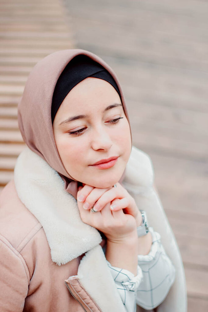 Muslim woman in beige coat and headscarf. Stylish Muslim woman in the park. A girl sitting on a bench - Photo, image