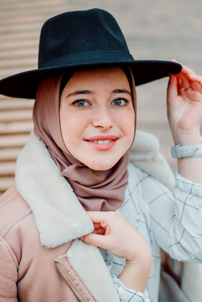 Muslim woman in beige coat and headscarf. Stylish Muslim woman in the park. Girl sitting on a bench wearing a black hat - Foto, afbeelding