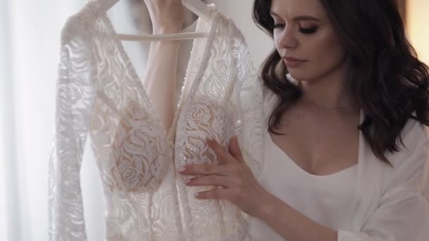 Noiva em lingerie de pé com seu vestido de noiva em casa perto da janela, vestido boudoir branco e véu - Filmagem, Vídeo