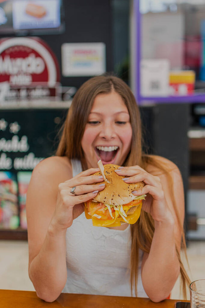 Una hermosa joven rubia está comiendo una hamburguesa con queso cheddar. Ella sonríe y es feliz. - Foto, imagen