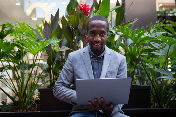 Αφρικανός επιχειρηματίας που κάθεται σε ένα παγκάκι δουλεύει με το λάπτοπ του. Επιτυχημένο άτομο με έμπιστο βλέμμα χαμογελά. Επιχειρηματίας εργάζεται σε εξωτερικούς χώρους - Φωτογραφία, εικόνα