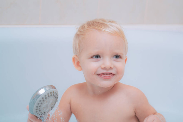 Funny cute baby is swimming in foam in the bath. Cute little baby boy taking bath playing with foam and duck toys in a white sunny bathroom. - Photo, image