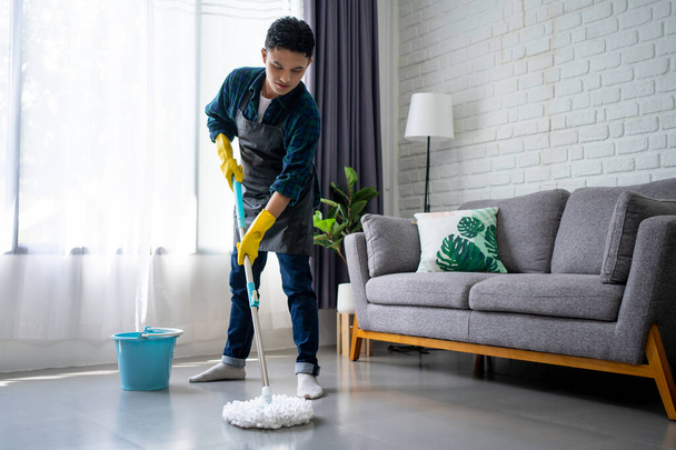 Handsome asian man wearing apron cleaning floor at home. Guy washing floor with mopping stick and bucket in living room. - 写真・画像