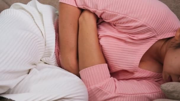 Young woman with arms on her stomach and stooping with severe abdominal pain, menstrual cramps or food poisoning is lying on the sofa at home. - Felvétel, videó