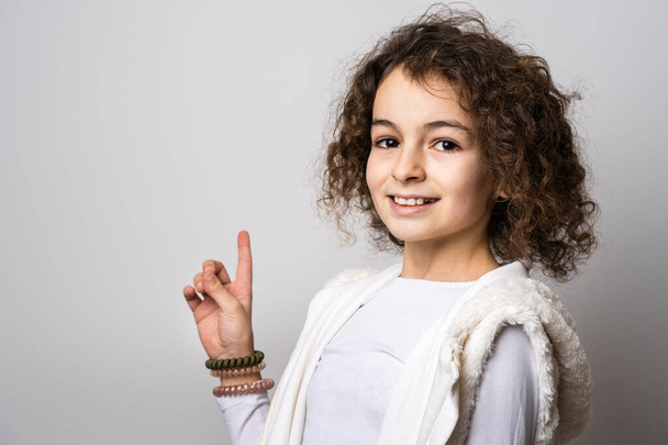 Одна маленькая кавказская девочка 10 лет, с кудрявыми волосами, портрет спереди, стоящий перед белым фоном, смотрящий в камеру, указывая пальцем, чтобы скопировать пространство, улыбаясь счастливой и радостной - Фото, изображение