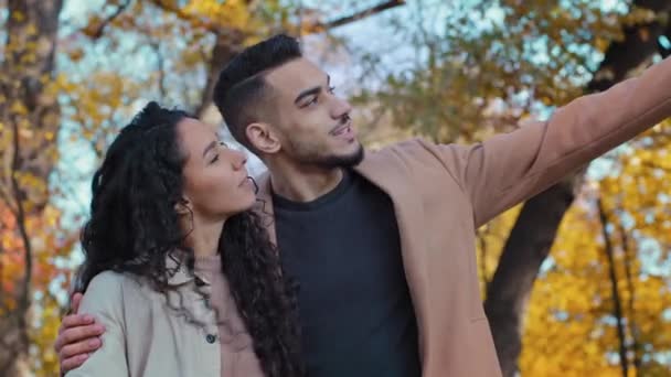 Happy hispanic young couple hugs in autumn park joyfully looking into distance enamored guy and girl cute communicate woman looks upward showing on sky outdoors man point finger side thinking dreamily - Filmati, video