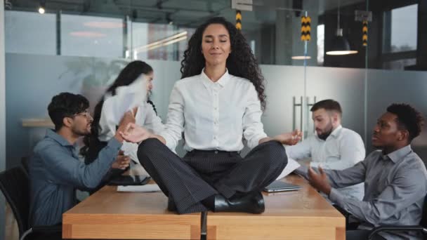 Young hispanic woman sitting on table in lotus position female leader meditating in office businesswoman feels calmness and serenity annoyed angry colleagues throw documents patience workplace concept - Filmagem, Vídeo