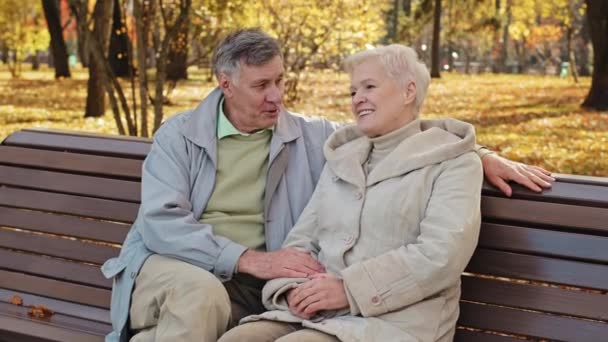 Serene caucasian older married couple rest sitting on bench in autumn park enjoy pleasant conversation outdoors man hugs beloved woman positive retirees enjoy life spends time together share good news - Video