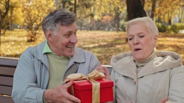 Mature man gives gift to beloved wife on birthday elderly woman happily laughs positive married couple celebrating anniversary unexpected surprise excited lady middle aged receive wrapped festive box - Filmati, video