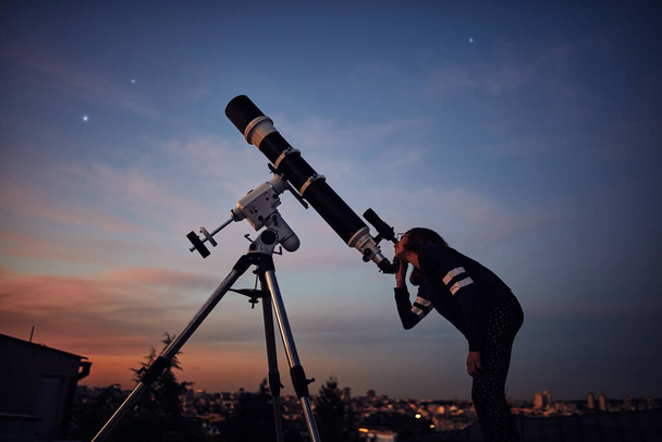 Девушка с астрономическим телескопом, смотрящая под сумеречным небом. - Фото, изображение