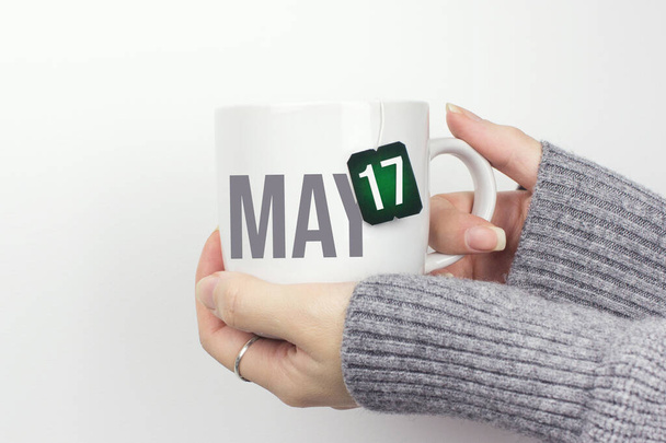 May 17th. Day 17 of month, Calendar date. Closeup of female hands in grey sweater holding cup of tea with month and calendar date on teabag label. Spring month, day of the year concept - Foto, Imagem