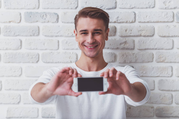 Mooie jonge man in casual wear met de telefoon in hand op de achtergrond witte bakstenen muur - Foto, afbeelding