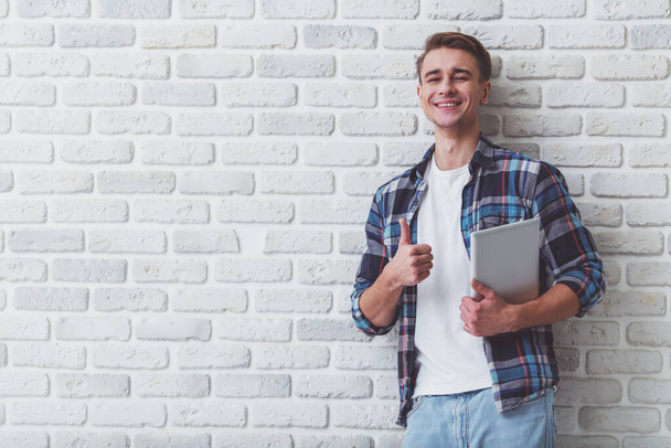 A beautiful young man, standing on a background of white brick wall with a tablet in hand - Photo, image