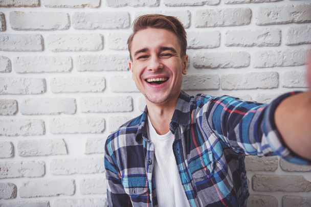 Όμορφη νεαρός άνδρας στο casual ρούχα κάνει selfie σε ένα φόντο λευκό τοίχο - Φωτογραφία, εικόνα