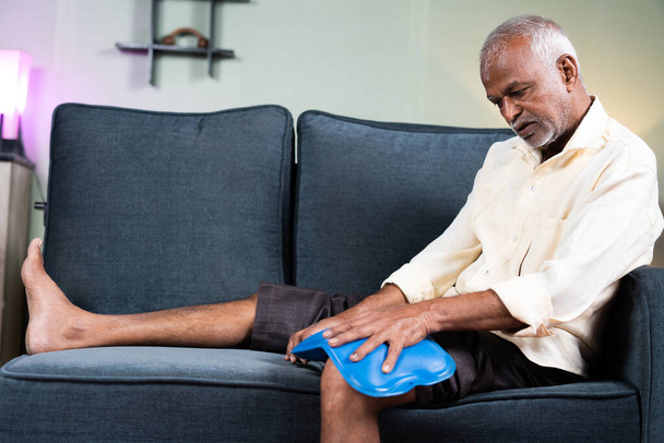 senior man using Hot water massage bag for knee joint pain relief while sitting on sofa at home - concept of natural hot water treatment, muscle relaxtion and osteoarthritis. - Photo, Image