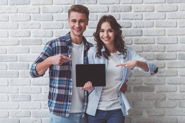 Happy young couple pointing finger at a blank black screen tablet on a brick wall background - Foto, afbeelding