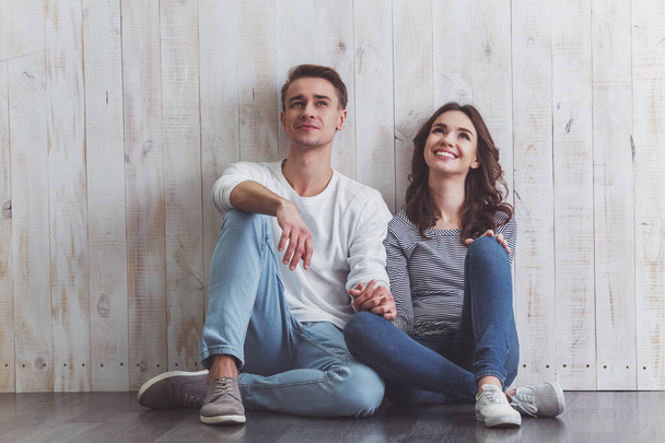Beautiful young couple sitting on the wooden floor, smiling in everyday clothes at home - Photo, Image
