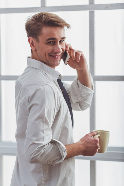 Beautiful young man in unbuttoned white shirt, talking on the phone, hold a cup in hand at home - Photo, Image