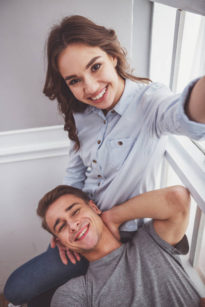 Νεαρό ζευγάρι χαρούμενος σε casual ρούχα, λαμβάνοντας selfie, που κάθεται στο περβάζι στο σπίτι - Φωτογραφία, εικόνα
