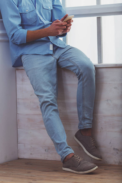 Beautiful young man in the blue shirt and light jeans, using smartphone, sitting on the windowsill at home - 写真・画像