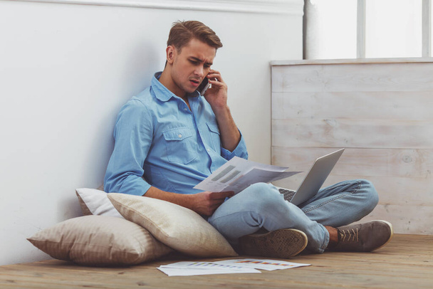 Young handsome man in the blue shirt and jeans, talking smartphone using laptop, sitting on cushions on the floor, next are graphics, sitting on the floor at home - Foto, Bild
