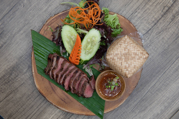 Overhead view of delicately decorated timber plate of nua yang Thai meal consisting of sliced seasoned steak, basekt of rice, and vegetable salad. - Foto, Bild