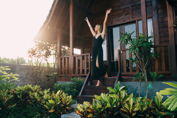 Full body of happy lady standing on wooden stairs of tropical house with raised arms and admiring view of nature - Fotó, kép