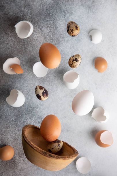 Ιπτάμενα αυγά κοτόπουλου και ορτυκιού και τσόφλια αυγού σε γκρι φόντο. Διατροφή ανύψωσης, χαρούμενη ιδέα του Πάσχα. - Φωτογραφία, εικόνα