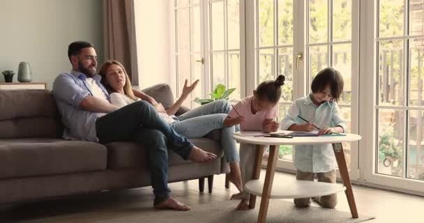 Loving couple talk on couch while little kids coloring pictures - Felvétel, videó