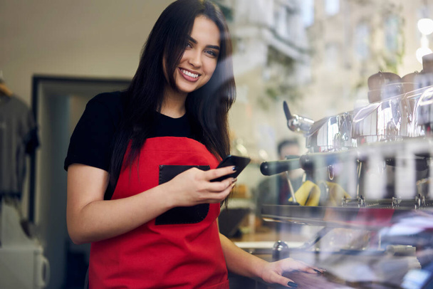 Prosperous brunette woman artist in apron using mobile phone while having break on job in cafeteria, 20s smiling female manager of coffee shop holding smartphone share content about job in blog - Foto, Imagen