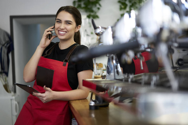 Smiling brunette woman waitress in uniform enjoying conversation on mobile phone making banking at work, prosperous female owner of coffee house calling to operator making order for retail business - Fotografie, Obrázek