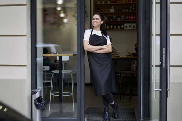 Cheerful brunette caucasian owner of coffee shop in apron standing at entrance greeting visitors and cliently hospitably, 20s smiling female barista in uniform opening cafeteria in morning - Valokuva, kuva