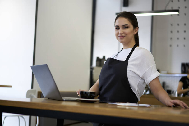 Half length portrait of 20s prosperous female employee of coffee shop in apron looking at camera checking business mails on laptop computer, millennial woman barista using technology for accountings - Foto, imagen